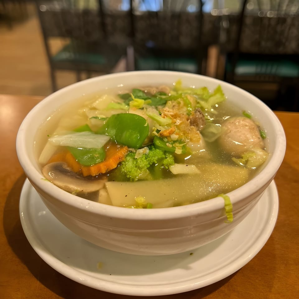 A white bowl of clear Thai soup with vegetables, mushrooms, and meatballs on a wooden table.