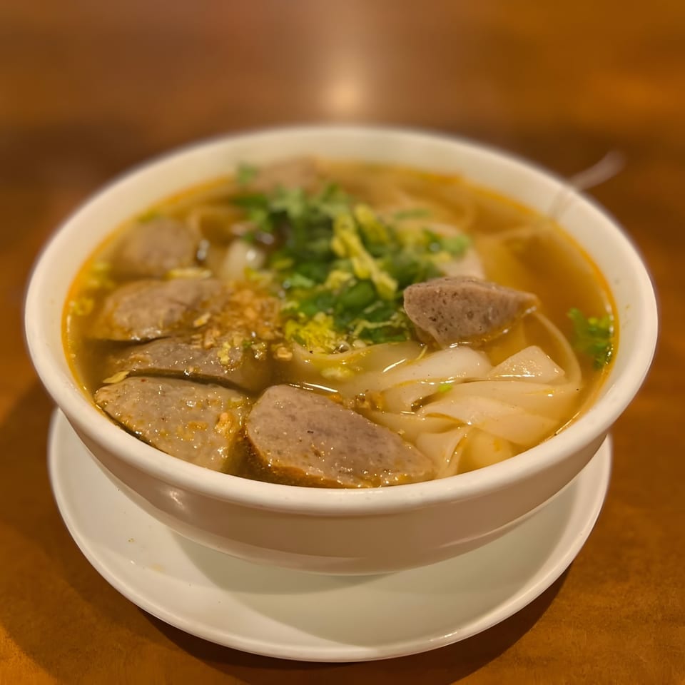 Thai noodle soup with sliced meatballs, wide rice noodles, and fresh herbs in a white bowl.